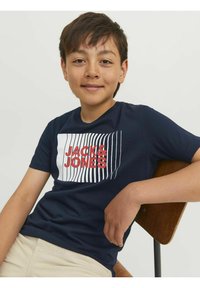Tumši zila kokvilnas tee ar grafiku, kurā redzams sarkans teksts "JACK&JONES" un vertikālas baltas līnijas. Modelis sēž uz koka krēsla.