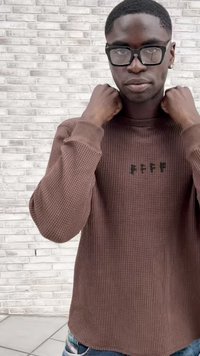 Brown waffle-knit sweater with long sleeves, featuring a small green text detail on the chest and a rounded hem. Modest, textured fabric.