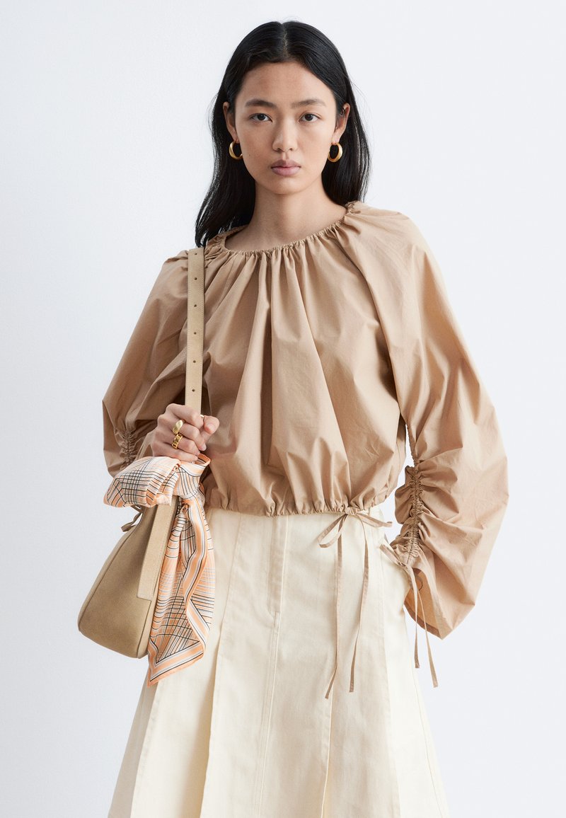Woman with long black hair wears beige puff-sleeve blouse and cream skirt, holding tan handbag with patterned scarf tied to strap.