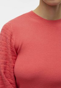 Koralinė megztinio su glotniuoju apykaklės ir tekstūrine nėrinių raštu ant rankovių. Minkšta medžiaga ir prigludęs dizainas. Be rašto kūno dalis.