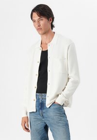 Weiße Knopfleiste-Cardigan aus weichem Material, mit Kragen, Brusttasche und goldenen Knöpfen, kombiniert mit einem schwarzen Tanktop und blauen Jeans.