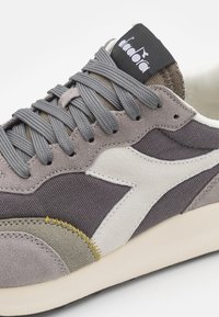 Sneaker grigio e beige realizzato in suede e tela, con lacci grigi, un logo bianco e dettagli in cucitura gialla lungo la suola.
