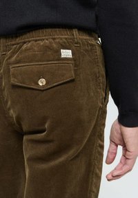 Pantalons en velours côtelé marron avec une texture à côtes prononcées, une seule poche arrière avec fermeture à bouton et une petite étiquette sur la poche.