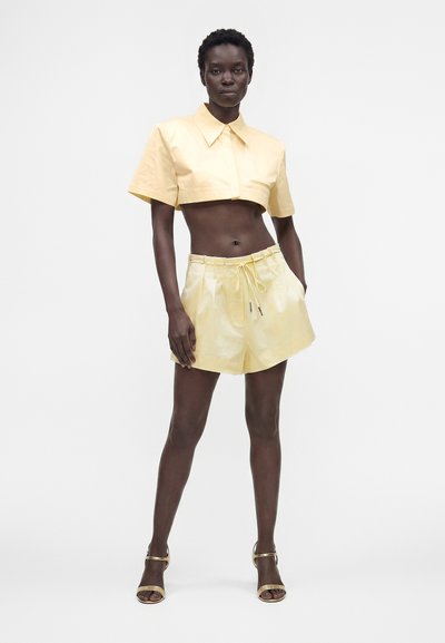 Femme debout portant un haut court à manches courtes jaune pâle, un short taille haute avec cordon et des talons ouverts assortis sur fond blanc.