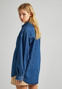 Camicia di denim blu scuro con maniche lunghe, parte anteriore con bottoni e taschino sul petto. Presenta un orlo curvo e una vestibilità rilassata.