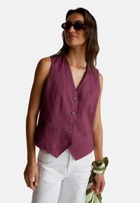 Kobieta w okularach typu tortoise, w bezrękawnikowej, śliwkowej kamizelce z guzikami, białych spodniach, trzymająca zielono-kremowy w paski szalik.