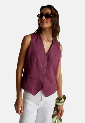 Donna che indossa occhiali da sole tartarugati, un gilet senza maniche color prugna con bottoni, pantaloni bianchi, mentre tiene una sciarpa a righe verde e crema.
