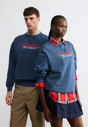 Two models stand side by side wearing matching blue sweatshirts with "Tommy Los Angeles" text, one paired with plaid shirt and skirt, the other with beige pants.