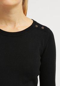 Suéter de punto negro con un cuello acanalado y dos botones decorativos en el hombro. Textura suave y diseño ajustado para mayor comodidad.