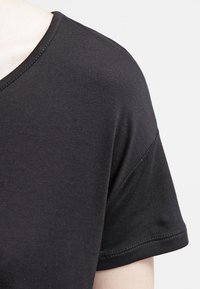 Närbild av en person som bär en enkel svart T-shirt med kort ärm mot en neutral ljus bakgrund.