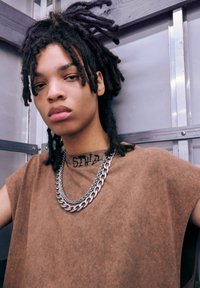 T-shirt oversized marron avec des manches roulées, collier en chaîne argentée épais, et un tatouage calligraphié visible sur la clavicule. Dreadlocks coiffés vers le haut.