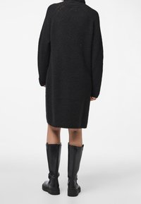 Personne portant une robe-pull noire longueur genou et des bottes en cuir noires hauteur genou, debout devant un fond blanc uni.