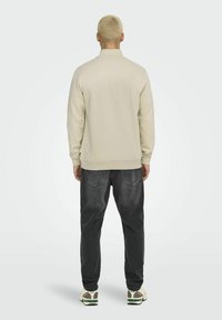 Sweatshirt beige avec un col montant, des poignets et un ourlet côtelés. Porté avec un jean gris foncé à coupe ample et des baskets blanches avec des accents verts.
