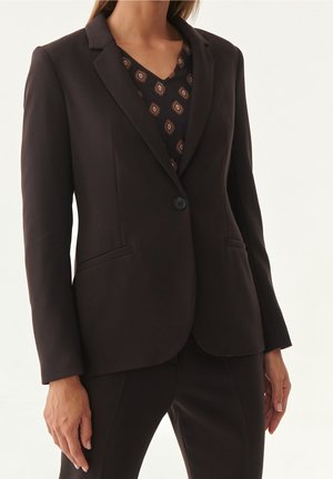 Frau trägt einen schwarzen, maßgeschneiderten Blazer mit einem Knopf über einer gemusterten Bluse mit V-Ausschnitt und passenden schwarzen Hosen.