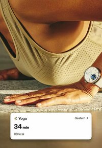 Helles beige Sportoberteil mit durchbrochener Gestaltung, gebräunte Haut und eine silberne Uhr am Handgelenk. Text auf dem Bildschirm zeigt Yoga-Dauer und verbrannte Kalorien.