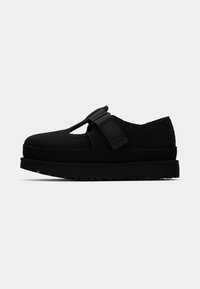 Zwarte platte schoenen van een zacht materiaal, met een enkele verstelbare band, dikke rubberen zool en een ronde neus.
