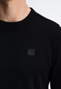 Schwarzer Strickpullover mit Rundhalsausschnitt und kleinem schwarzen Logopatch auf der Brust. Der Stoff wirkt weich und hat eine strukturierte Oberfläche.