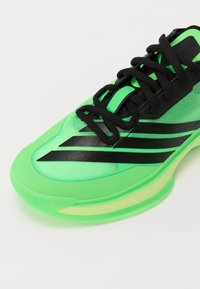 Pantofii atletici verzi cu dungi negre, partea superioară din plasă și talpă translucidă. Au un vârf rotunjit și detalii de șireturi negre.