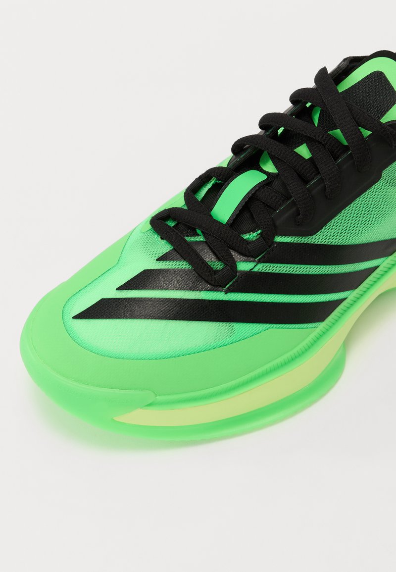 Zapatilla deportiva verde con rayas negras, parte superior de malla y suela translúcida. Tiene una punta redondeada y detalles en negro en los cordones.