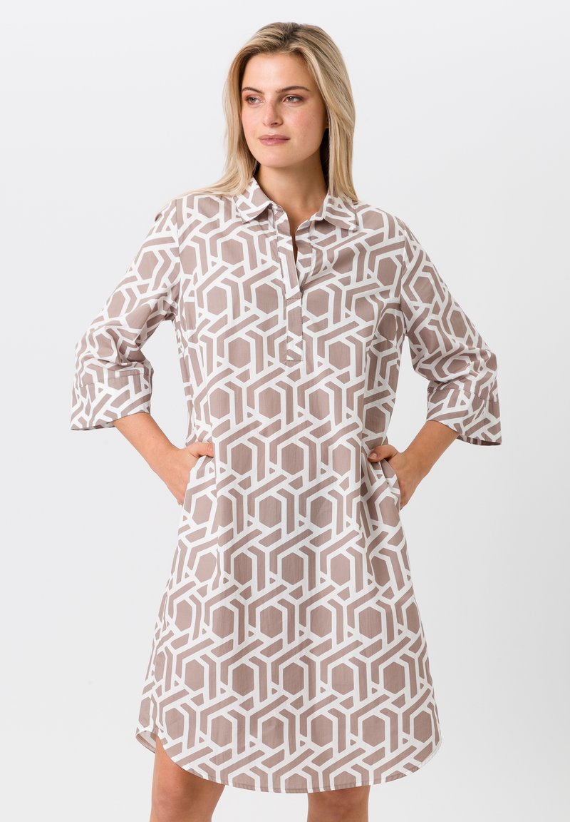 Robe chemise légère en beige avec un motif géométrique blanc. Dotée d'un col, d'une fermeture à boutons sur le devant et de manches 3/4 retroussées.