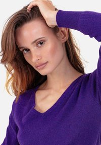 Lila V-Ausschnitt-Pullover aus weichem Material mit figurbetontem Design. Mit gerippten Bündchen und glatter Struktur. Modell posiert mit Hand im Haar.