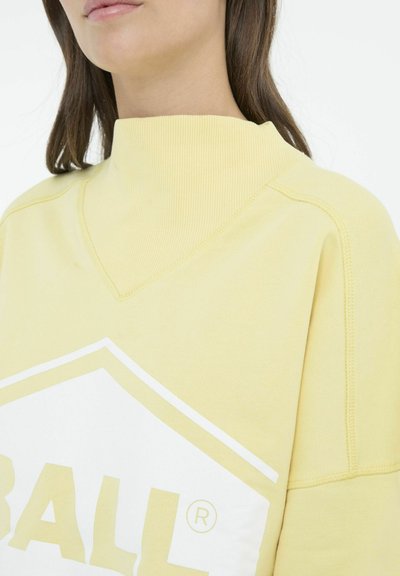 Gul sweatshirt med en ribbet høj krave, der har et hvidt grafisk logo. Stoffet ser blødt ud, med synlige syninger.