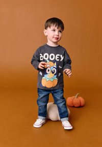 Graues Langarmshirt mit dem Schriftzug "BOOEY" und einem Cartoon-Hund in einem Kürbiskostüm. Kombiniert mit blauen Jeans und weißen Sneakers.