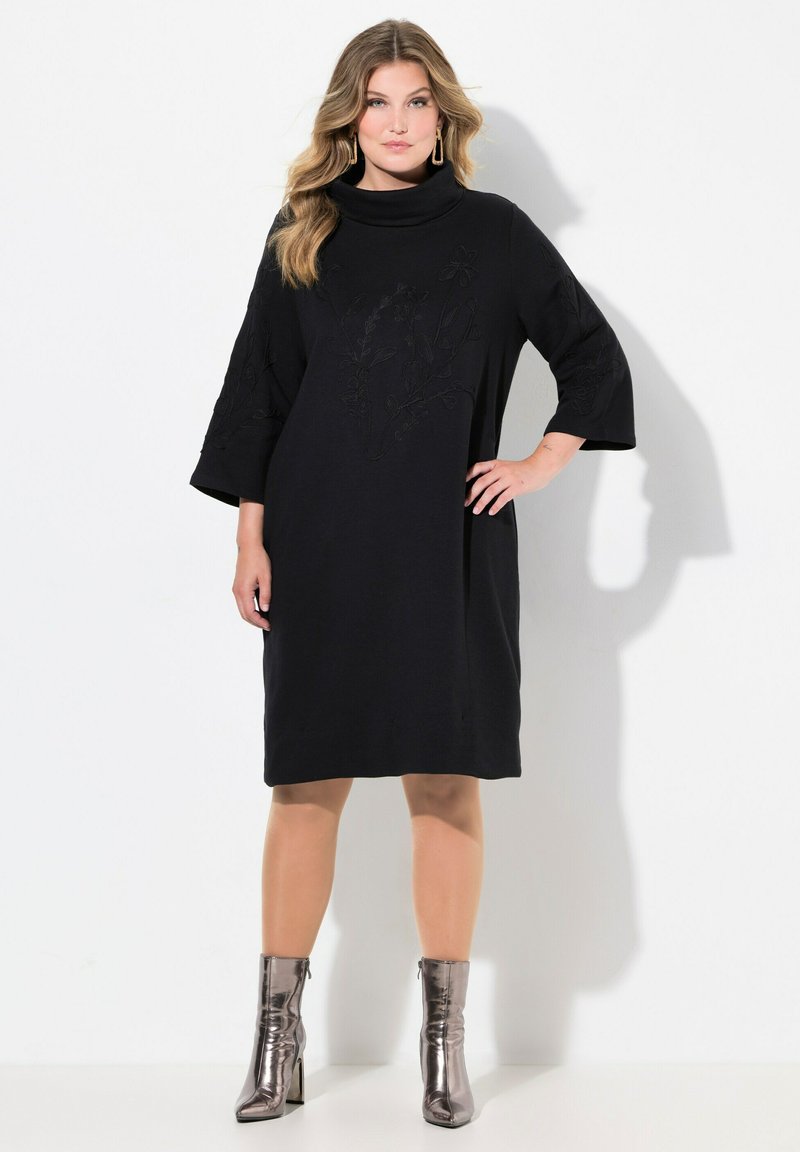 Vestido negro con cuello alto, mangas tres cuartos y bordado floral. Combinado con botines metálicos, creando un contraste en textura.