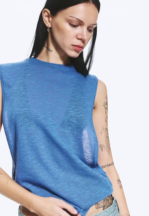 Jeune femme aux longs cheveux noirs portant un haut en maille bleu sans manches, montrant des tatouages sur les bras et le torse, regardant vers le bas.