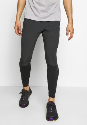 Partie inférieure du corps d'une personne portant un pantalon de sport noir fuselé et des chaussures de course noires avec des touches violettes et jaunes.