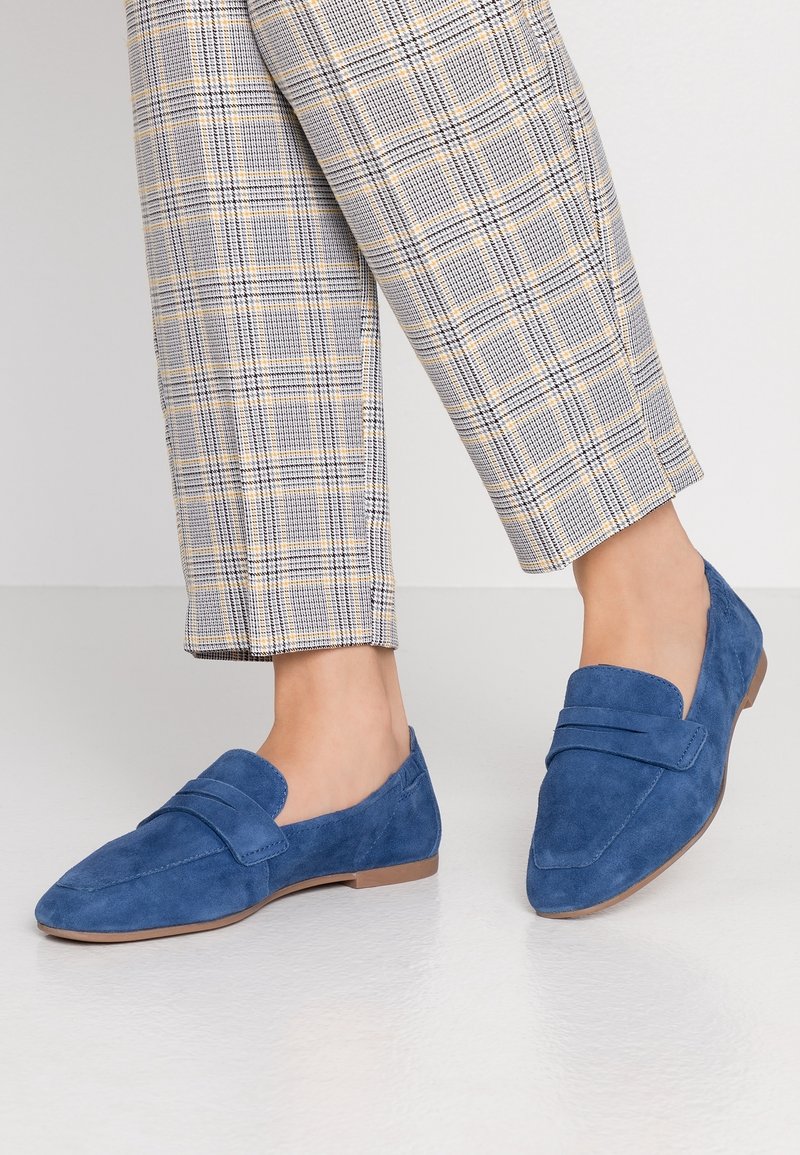 Mocassins en daim bleu avec une semelle plate, dotés d'une lanière à l'avant et d'un design à bout pointu. Portés avec un pantalon à motif écossais.
