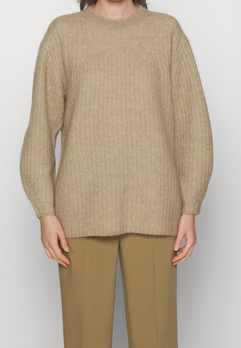 Personne portant un pull beige en maille côtelée à manches longues et col rond, associé à un pantalon couleur chameau.