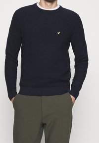 Homme portant un pull texturé bleu marine avec un petit logo d'oiseau jaune sur la poitrine et un pantalon vert olive, mains dans les poches.