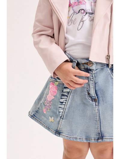 Παιδί που φοράει ανοιχτό ροζ σακάκι και τζιν φούστα με κεντήματα από λουλούδια και πεταλούδες, κρατώντας το τελείωμα της φούστας.