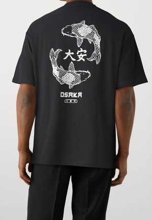 Czarny t-shirt z białą grafiką dwóch ryb koi krążących wokół japońskich znaków i słowa „Osaka” na plecach, noszony przez osobę.