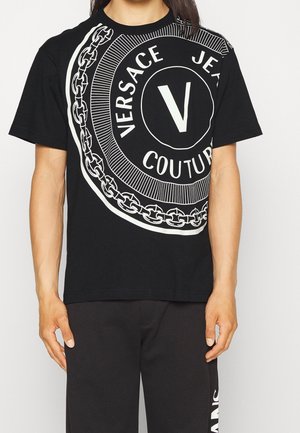 Schwarzes Baumwoll-T-Shirt mit einem großen weißen runden Logodesign, das "Versace Jeans Couture" und ein Kettenmuster rund um den Kreis zeigt.