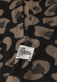 Leopardmönstrad tyg i svart och beige med en texturerad yta. Etiketten lyder: "HITTA PARADIS VAR DU ÄN GÅR" i svart text på vit bakgrund.