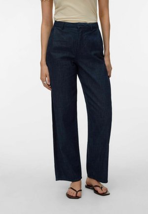 Persoon die donkerblauwe wijde jeans, beige top met korte mouwen, zilveren ring en bruine sandalen met vierkante neus draagt, tegen een effen achtergrond.