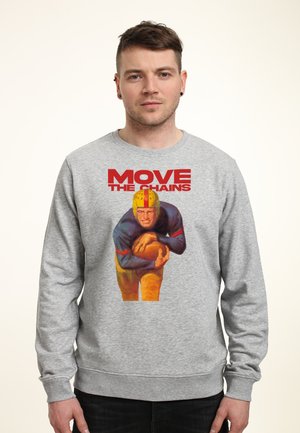 Mężczyzna w szarej bluzie z nadrukiem vintage przedstawiającym futbolistę trzymającego piłkę oraz czerwonym napisem "MOVE THE CHAINS" na przodzie.