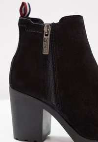 Botte cheville en daim noir avec talon bloc, fermeture éclair latérale étiquetée "Hilfiger Denim", et languette de tirage en tissu rouge, blanc et bleu à l'arrière.