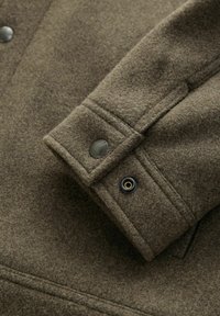 Brauner Wolljackenbund mit strukturiertem Finish, Druckknopf im Militärstil und verstärktem Nähdetail.
