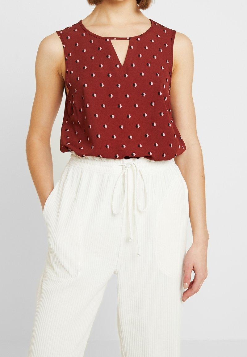 Haut bordeaux sans manches avec de petits motifs en losange et un décolleté en forme de goutte, associé à un pantalon blanc taille haute avec cordon de serrage.