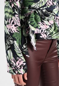 Blouse à motifs floraux avec des motifs verts, roses et noirs ; manches longues avec liens ; associé à un pantalon ajusté marron. Texture de tissu lisse.