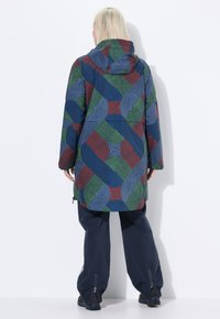 Osoba v kapucínovém kabátu s modrým, zeleným a červeným geometrickým vzorem, v kombinaci se volnými námořnickými kalhotami a černými botami, stojící proti zdi.