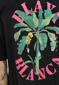 Sort t-shirt med et livligt grønt plantegrafik, med pink tekst der læser "PLAK BLANCA" og "SINCE MCMXC" omkring designet.