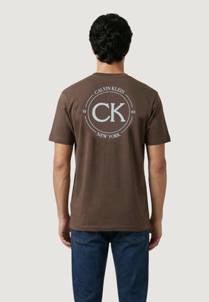 Uomo che indossa una maglietta marrone Calvin Klein con logo e scritta bianca sul retro, abbinata a jeans blu, in piedi di spalle.