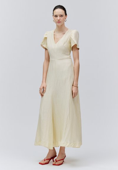 Frau steht in einem cremefarbenen Midi-Kleid mit V-Ausschnitt, aufgebauschten kurzen Ärmeln und roten Sandalen mit Absatz vor einem schlichten grauen Hintergrund.