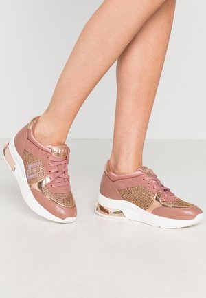 Jambes portant des baskets roses et or rose avec des semelles blanches, présentant des accents en mesh et métalliques sur un fond clair.