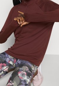 Bordeauxfarbenes Langarmoberteil mit einem halben Reißverschluss am Kragen. Kombiniert mit Leggings im Blumenmuster in verschiedenen Rosa- und Grautönen. Strukturierter Stoff sichtbar.
