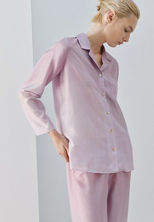 Kobieta o blond włosach związanych w niski kok, ubrana w jasnofioletową, długorękawową koszulę na guziki i pasujące spodnie, stojąca na tle jednolitego jasnego tła.